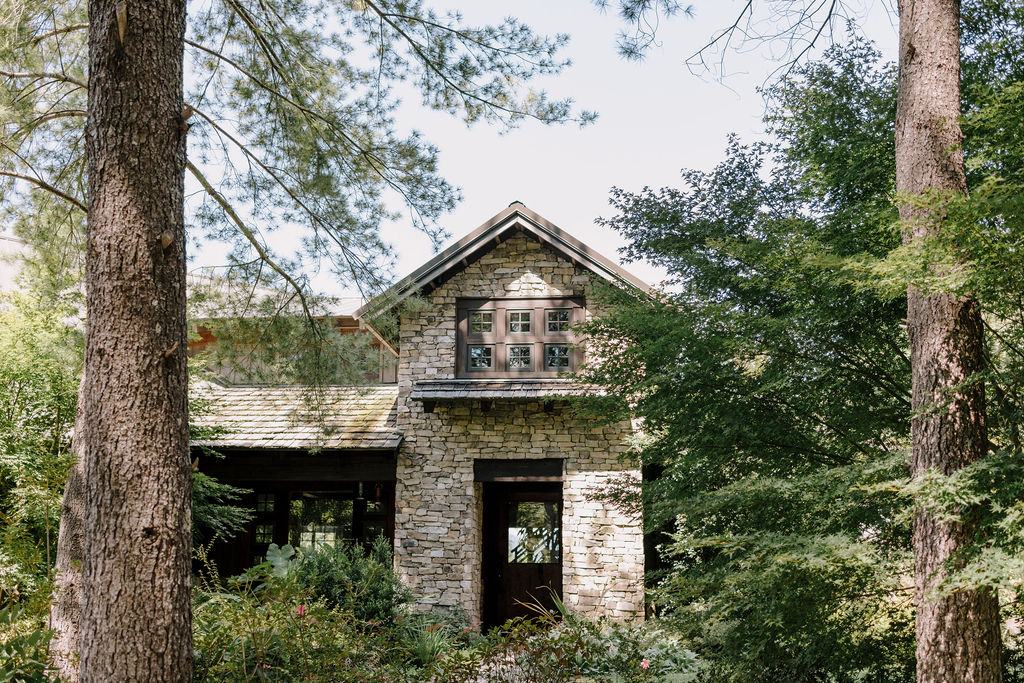 A medium-range, elevated photograph looking down upon the stone-faced, multi-level entrance of House Boheme. The structure is nestled within a lush, garden-like setting of mature pines and dense deciduous trees, with some early fall colors visible in the foliage.