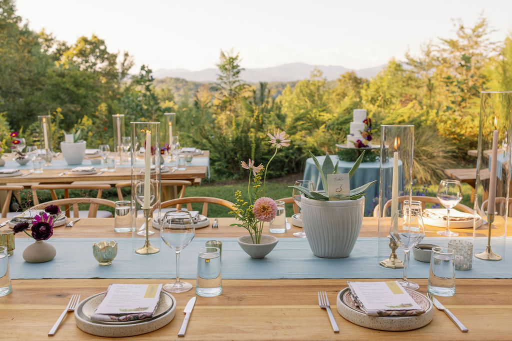 An outdoor wedding reception setup at golden hour with the Blue Ridge Mountains in the background. Long wooden dining tables are set with light blue runners, modern glass hurricane candles, small wildflower centerpieces, and a potted agave plant. Custom menu cards sit on speckled ceramic plates.