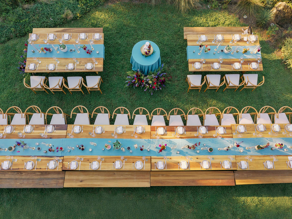 A symmetrical overhead drone photograph of an outdoor wedding reception on a green lawn. One long, continuous wooden table stretches across the bottom, balanced by two shorter rectangular tables at the top. A round, blue-clothed cake table sits centered between them. All tables feature matching light blue runners and minimal centerpieces.
