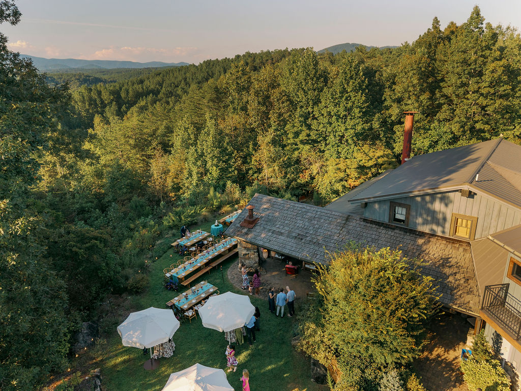 Wide-angle landscape photograph looking out over a reception area with long wooden tables and a view of the Blue Ridge Mountains at sunset. The setting features a dusty blue table runner, various-sized candle holders, and bud vases with mixed wildflowers. A small, simple wedding cake sits on a pedestal in the distance.