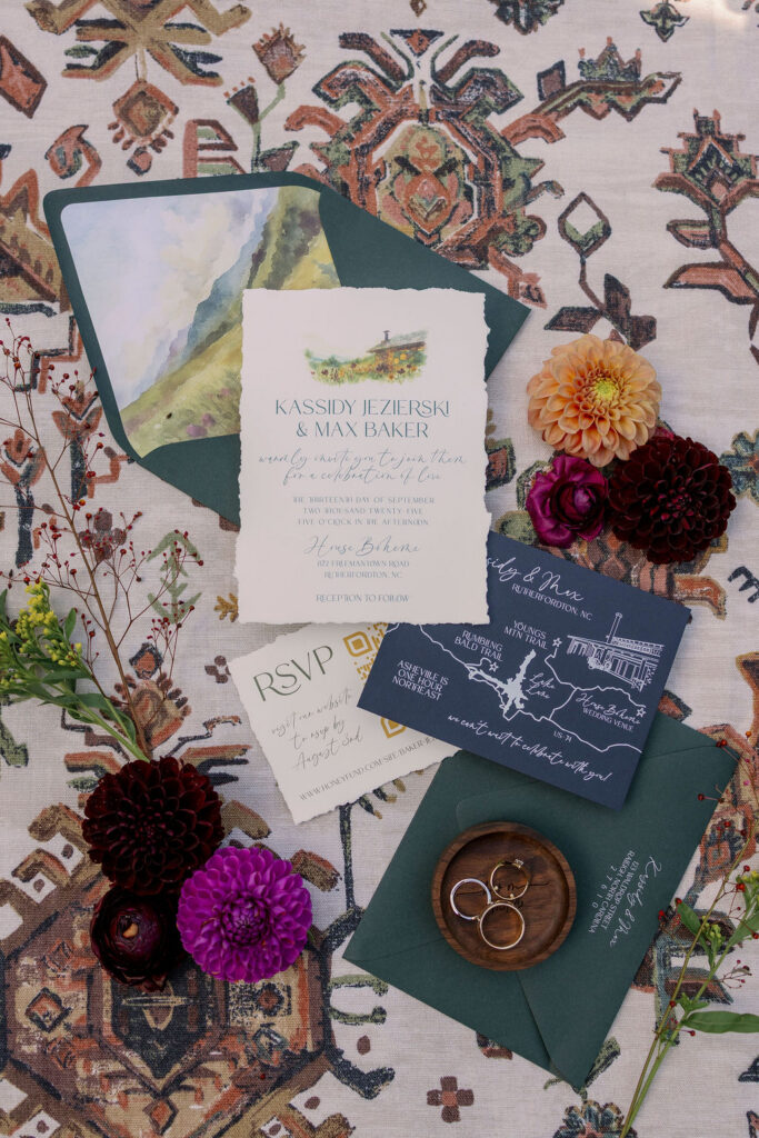 Flat lay photograph of Kassidy and Max's custom watercolor wedding invitation suite. The main invitation, with a deckled edge and illustration of the House Boheme venue, is centered. Surrounding it are emerald green envelopes with mountain-scape liners, an RSVP card with a QR code, and a dark blue map card with white line art details.
