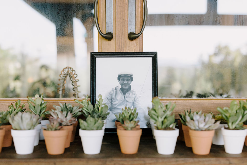 A framed black and white portrait photograph of the groom's late father is positioned centrally, honoring his memory. In front of the portrait, a long row of uniform, miniature terracotta-potted succulent favors is lined up, ready for guests to take home.