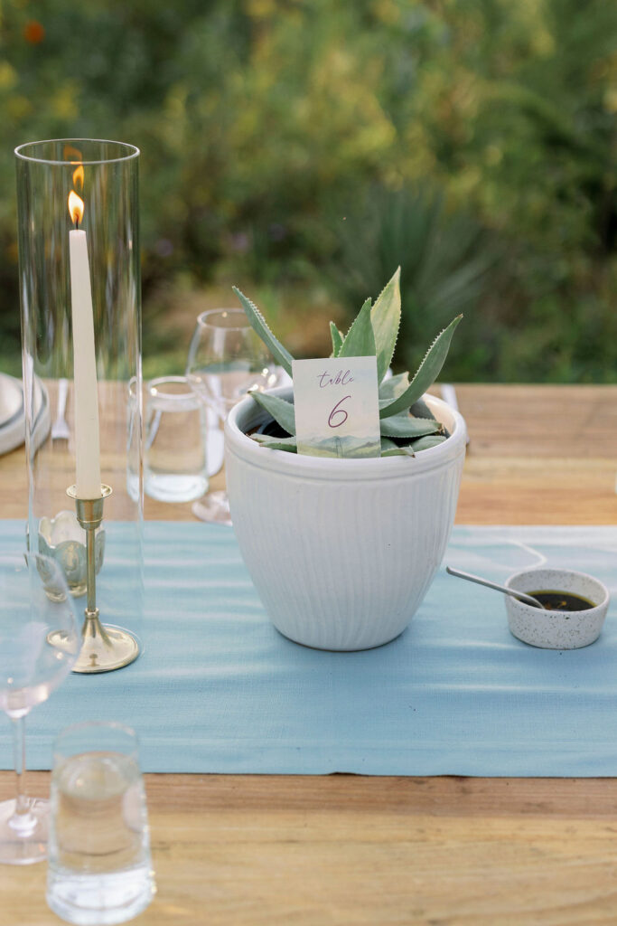 Elegant wedding tabletop decor featuring a custom watercolor table number card reading "Table 6," nestled inside a potted green agave plant. The setup rests on a light blue fabric runner alongside a tall, modern glass hurricane candle holder with a lit white taper candle.