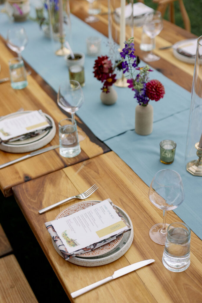 Angled close-up of an outdoor wedding reception table setting. A personalized menu card reading "Larry" with a yellow-gold accent border rests on a patterned salad plate stacked on a speckled ceramic charger. The wooden table is decorated with a light blue fabric runner, clear glassware, silver flatware, small green votive holders, and minimal concrete bud vases holding deep red and purple wildflowers.