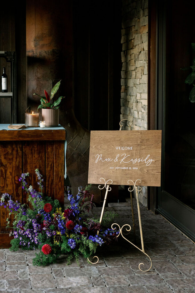 Medium shot of a wooden welcome sign for "Max & Kassidy" set on a gold-colored wire easel on a stone patio. A vibrant, bohemian floral arrangement with deep reds and purples sits at the base of the easel, next to the entrance of House Boheme.