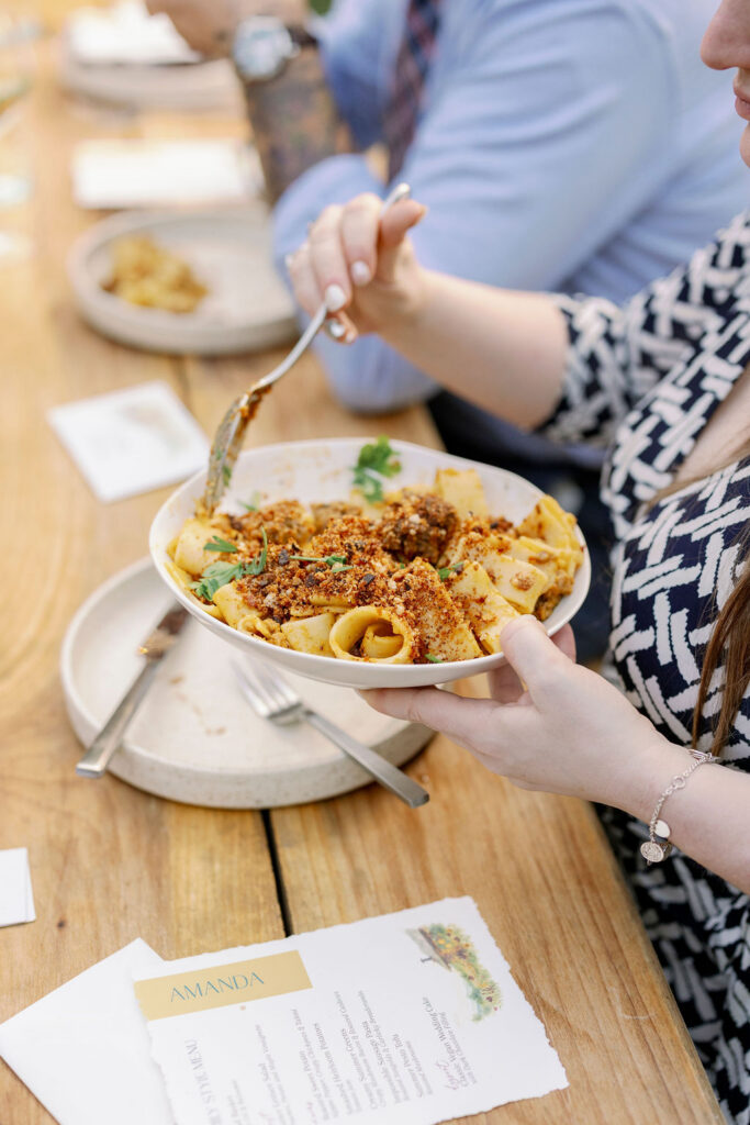 A wedding guest holds a white ceramic bowl filled with a family-style pasta dish topped with breadcrumbs and herbs over a wooden reception table. In the foreground, a personalized deckled-edge wedding menu card reads "Amanda" with a custom venue illustration.