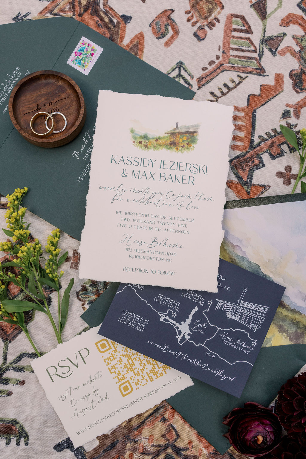 Flat lay photograph of Kassidy and Max's custom watercolor wedding invitation suite including their weddings rings in a custom wooden holder. The main invitation, with a deckled edge and illustration of the House Boheme venue, is centered. Surrounding it are emerald green envelopes with mountain-scape liners, an RSVP card, and a dark blue map card with white line art details.