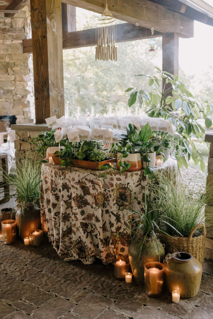 A bohemian-styled escort card display featuring individual seed packets personalized with guest names and table numbers. The packets are clipped to small transparent holders and nestled into a mix of live plants, creating a lush, organic wall of paper and greenery.