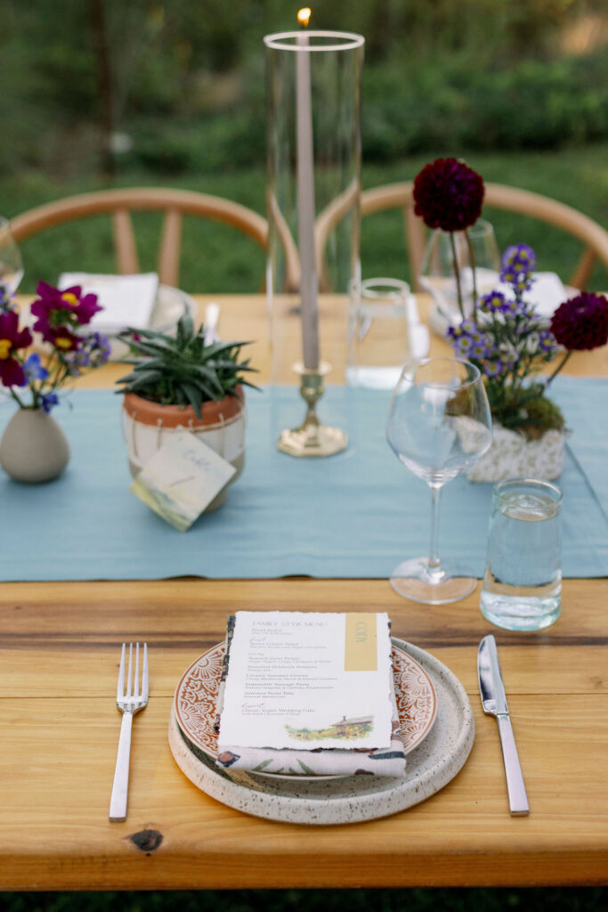 A beautifully styled wedding reception place setting at an outdoor wooden table. A patterned ceramic plate holds a custom family-style menu card reading "Cody," accented by a yellow-gold border. The table runner features a small potted succulent, a tall hurricane candle, and bud vases with deep purple and blue wildflowers.