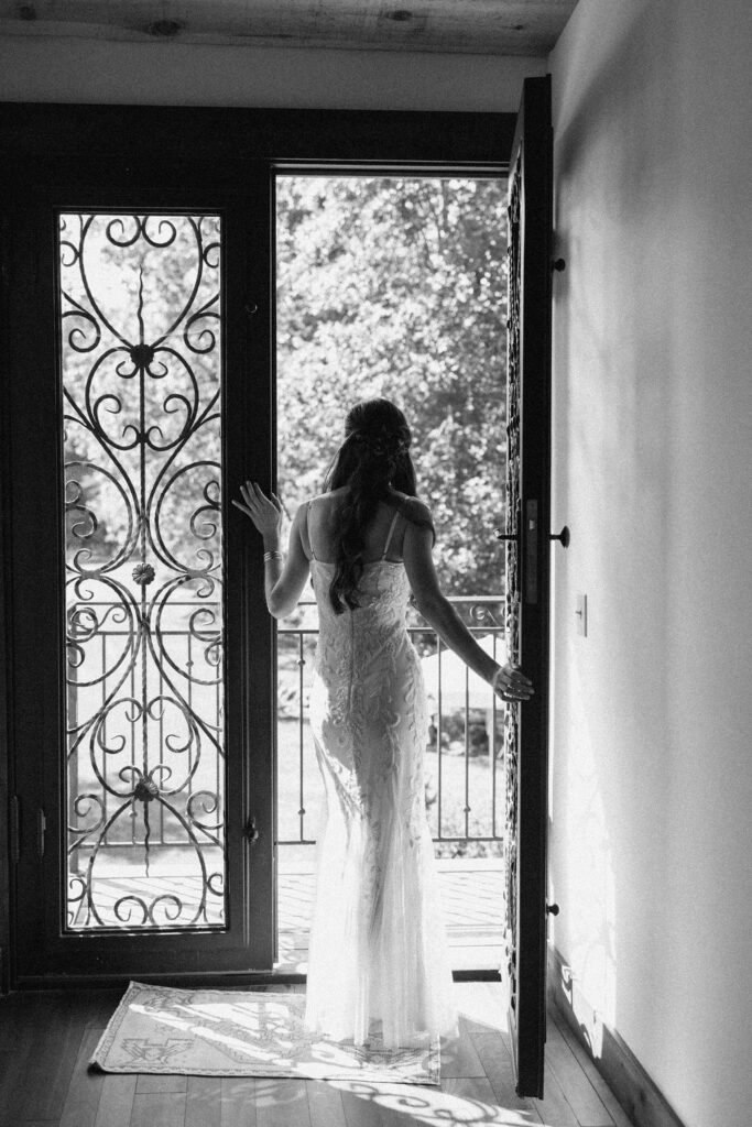 A black and white portrait of the bride, Kassidy, from behind. She stands at a large, iron-wrought glass door leading out to a balcony, wearing a floor-length wedding gown with floral beading. Her arms are raised as she looks out, with the light from outside casting reflections.