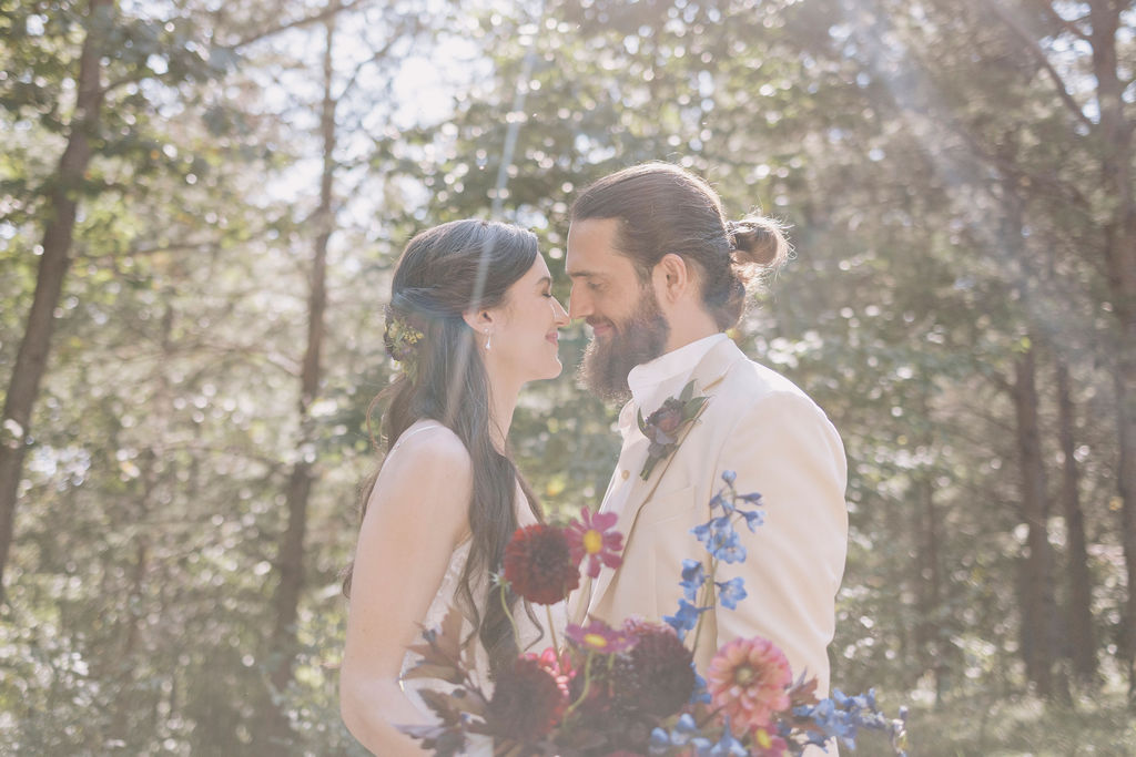Head-and-shoulders portrait of the bride, Kassidy, and groom, Max, standing close together in a wooded area with sunlight filtering through the trees. Kassidy smiles while looking down, and Max looks at her, as they hold a colorful dahlia and delphinium bouquet.