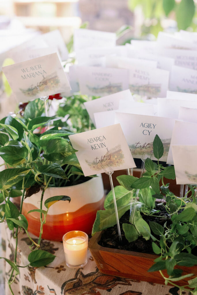 Close-up of a whimsical wedding seating chart display. Small, custom watercolor seed packets printed with guest names and table numbers are styled inside a rustic wooden planter box filled with lush, leafy green herbs. A small glowing votive candle sits on the patterned tablecloth nearby.